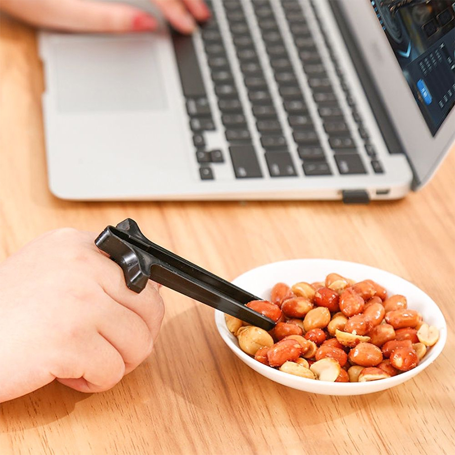 A hand using chopstick-style tongs to pick peanuts from a bowl while working on a laptop.