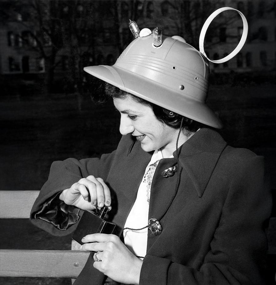 A woman wearing a helmet with attached wires, a circular antenna, and small light bulbs sits on a bench, smiling as she adjusts a small device in her hands.