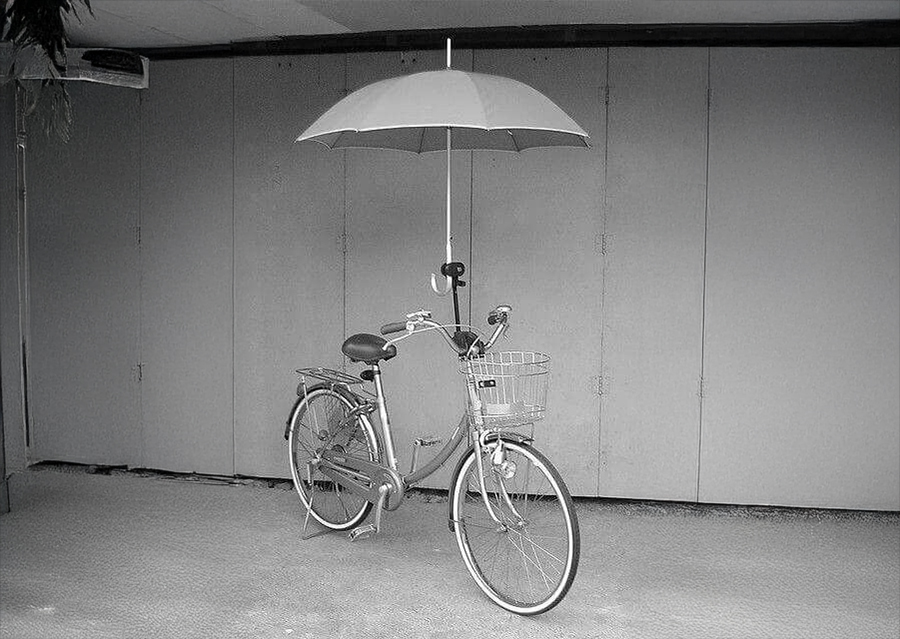 A bicycle with a large umbrella attached above the handlebars stands near a grey wall. The umbrella is open, providing coverage over the bike.