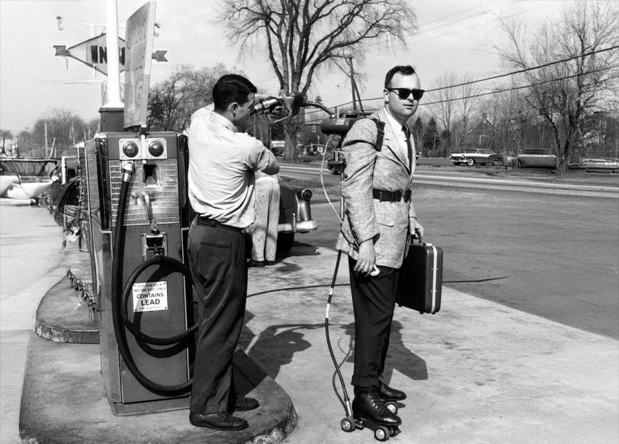 A man on roller skates, wearing a jetpack and holding a briefcase, stands at a gas station while another man fuels the jetpack. Vintage black and white photo with cars and trees in the background.