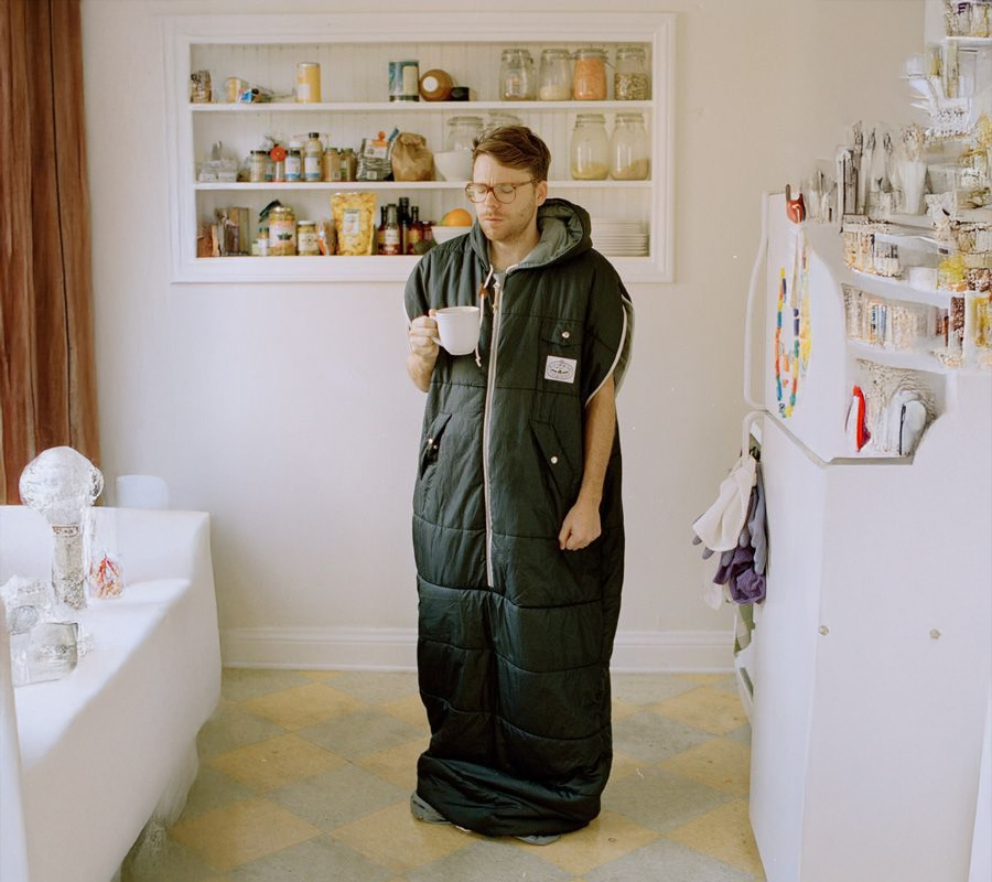 A person stands in a kitchen wearing a sleeping bag like a coat, holding a mug. Shelves with jars and containers are visible behind them.