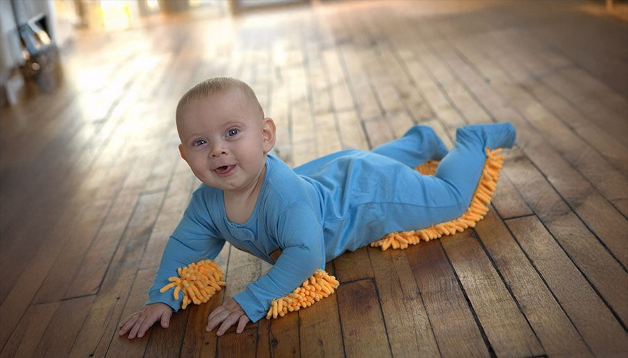 A smiling baby in a blue onesie with orange mop-like fringes lies on a wooden floor, appearing to help clean while crawling.