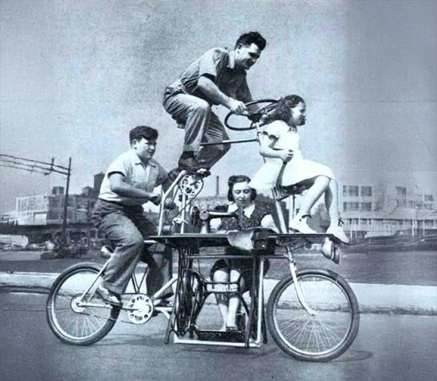 Four people ride a bicycle built around a sewing machine: one pedals, one sews, one perches on top, and a child sits on the back, with an urban landscape in the background.