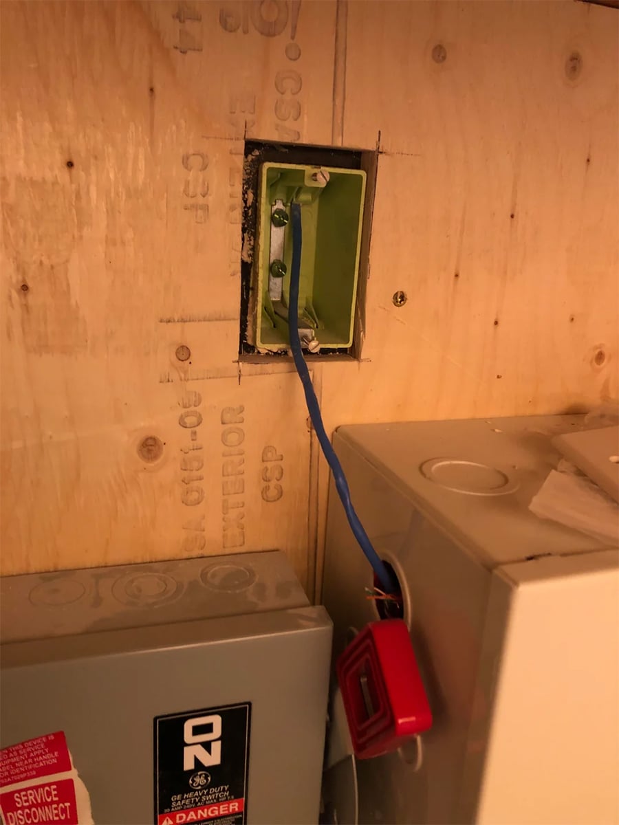 A green electrical box set into plywood, with an exposed blue wire extending out. Nearby are gray utility boxes, one labeled “ON,” and a small red plastic tool attached to a wire.