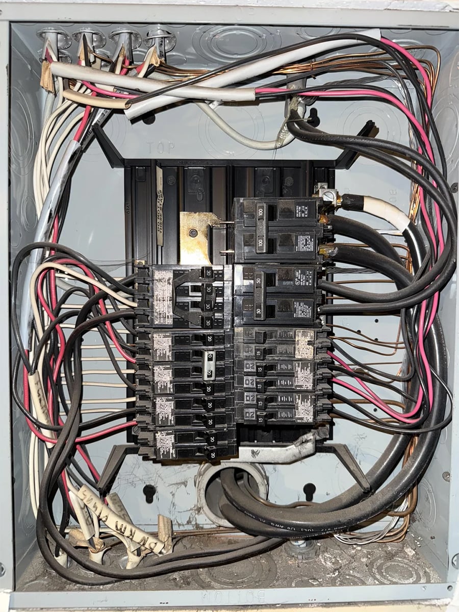 An open electrical breaker panel with multiple circuit breakers, black, white, and red wires connected, and various cables entering from the top and bottom. The panel is mounted on a wall and shows signs of dust and minor wear.