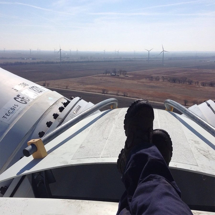 Person’s feet in boots rest on top of a wind turbine, overlooking a flat landscape with several other wind turbines in the distance under a clear sky.