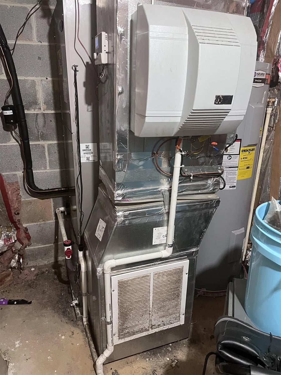 A home HVAC unit with attached humidifier in a basement, surrounded by pipes and ductwork. A water heater and concrete wall are visible in the background, with a blue plastic bin to the right.