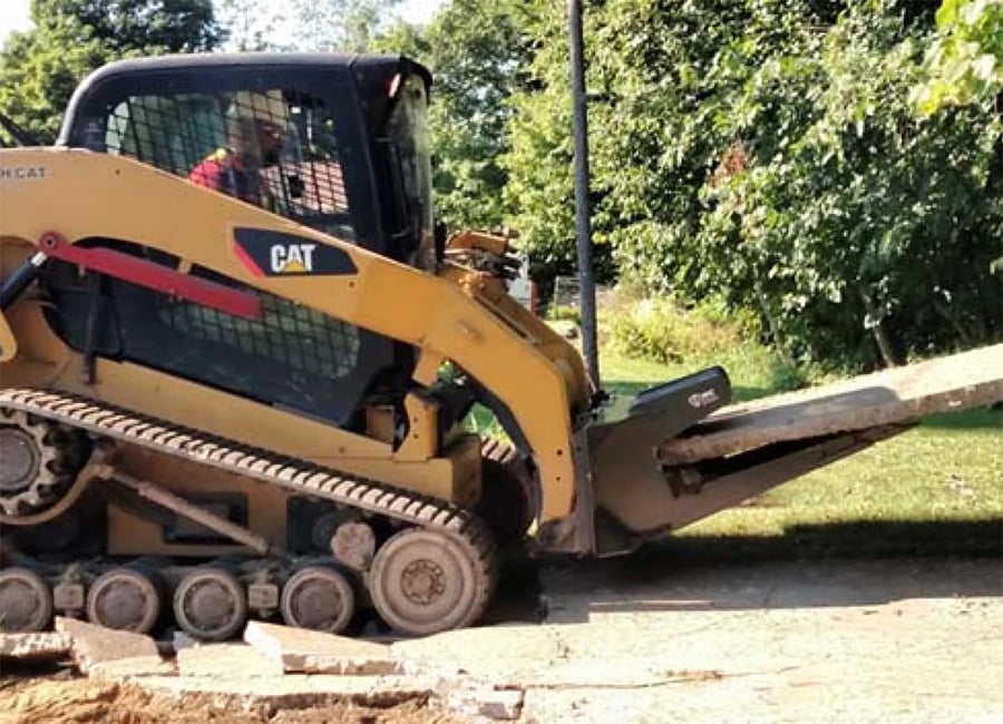 A person operates a yellow CAT skid-steer loader, lifting a large piece of broken concrete near a leafy, tree-lined area on a sunny day.