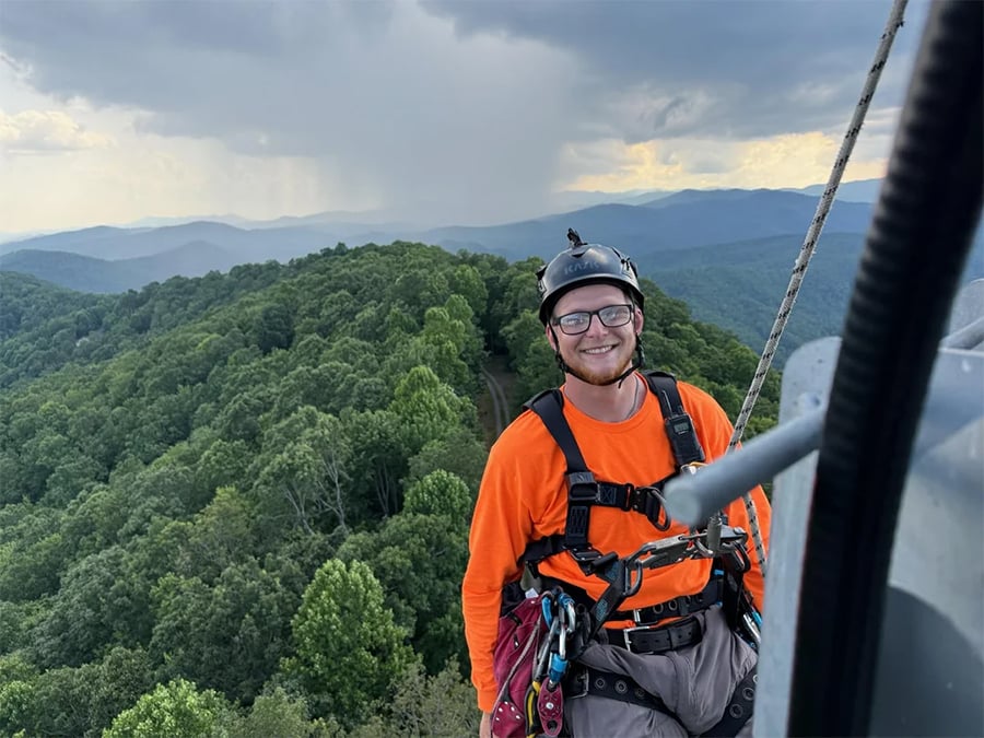 A person in safety gear and an orange shirt smiles while secured to a harness on a high structure above a forested mountain landscape with cloudy skies.