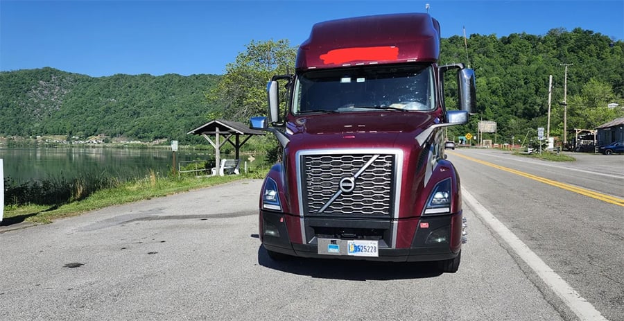 A maroon Volvo semi-truck is parked on the roadside near a lake, with green hills and a blue sky in the background.