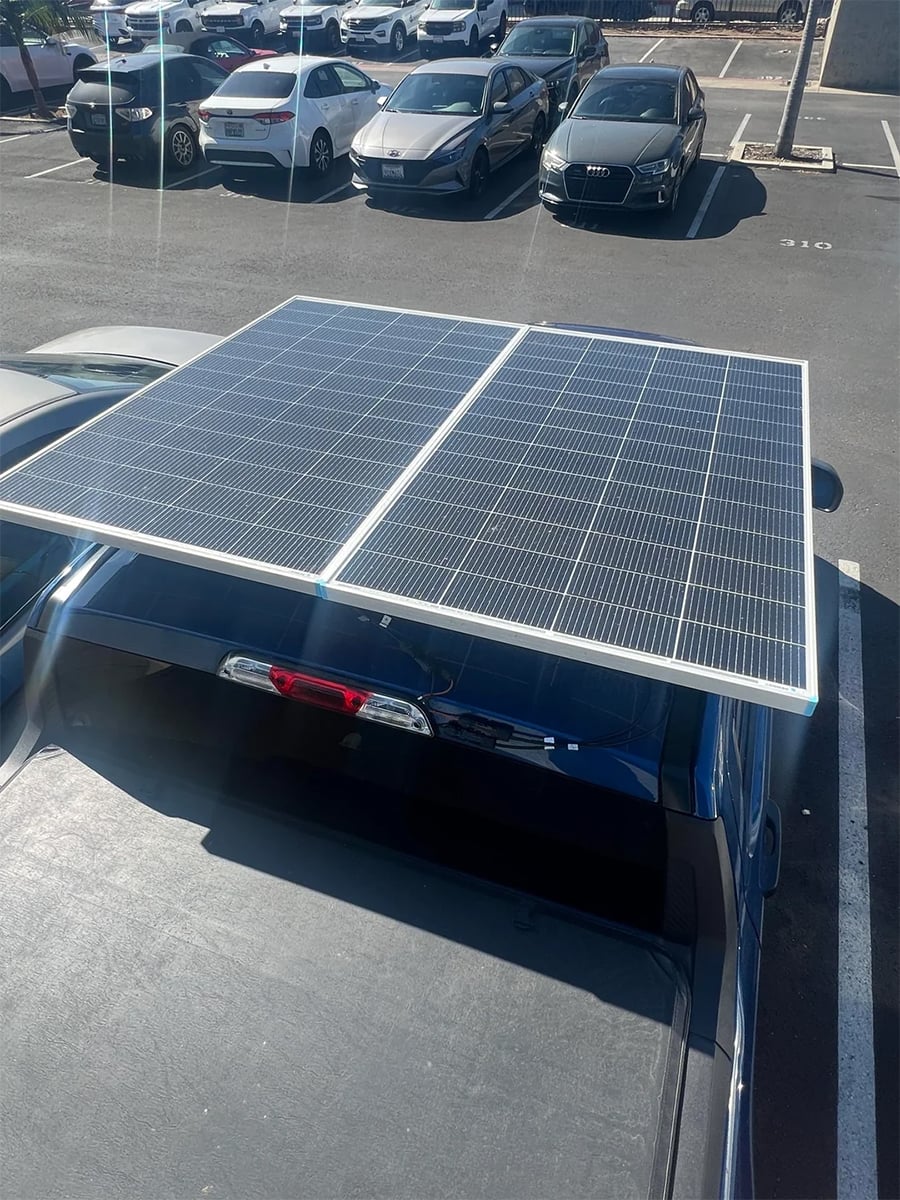 Two large solar panels are mounted on top of a truck parked in a parking lot, with several other cars visible in the background.
