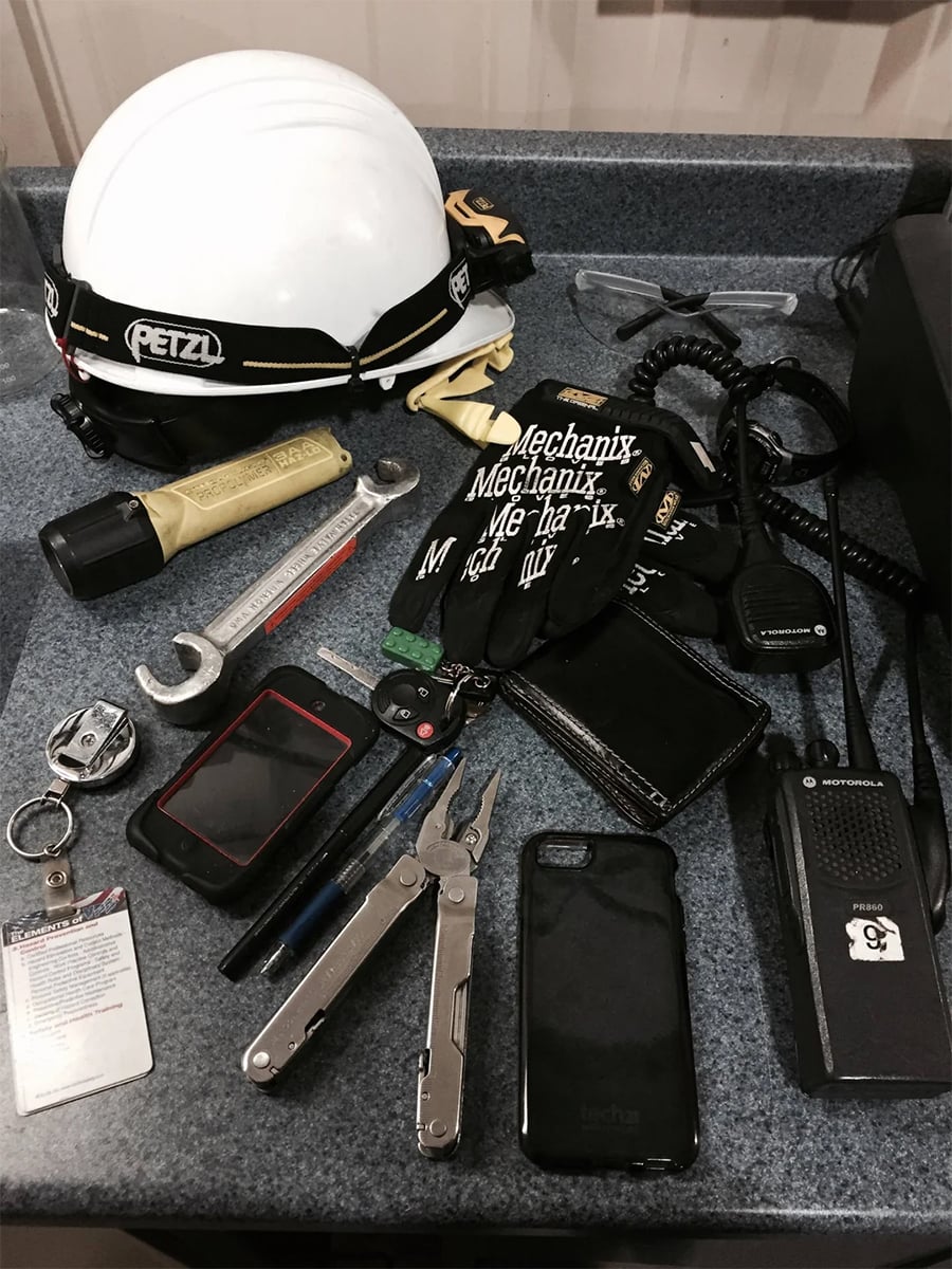 Various tools and gear on a counter, including a white safety helmet, gloves, flashlight, wrenches, multitool, pen, notepad, wallet, keys, ID badge, radio, and smartphone. Items are neatly arranged.