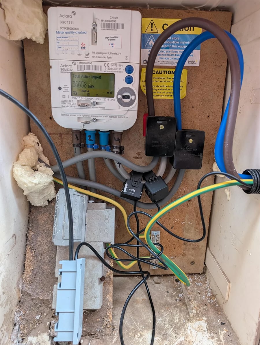 An open electric meter box shows wires connected to a smart meter, warning labels, and insulation foam on the left. Various cables are visible, with some connected to black blocks and a grey fuse holder is at the bottom left.