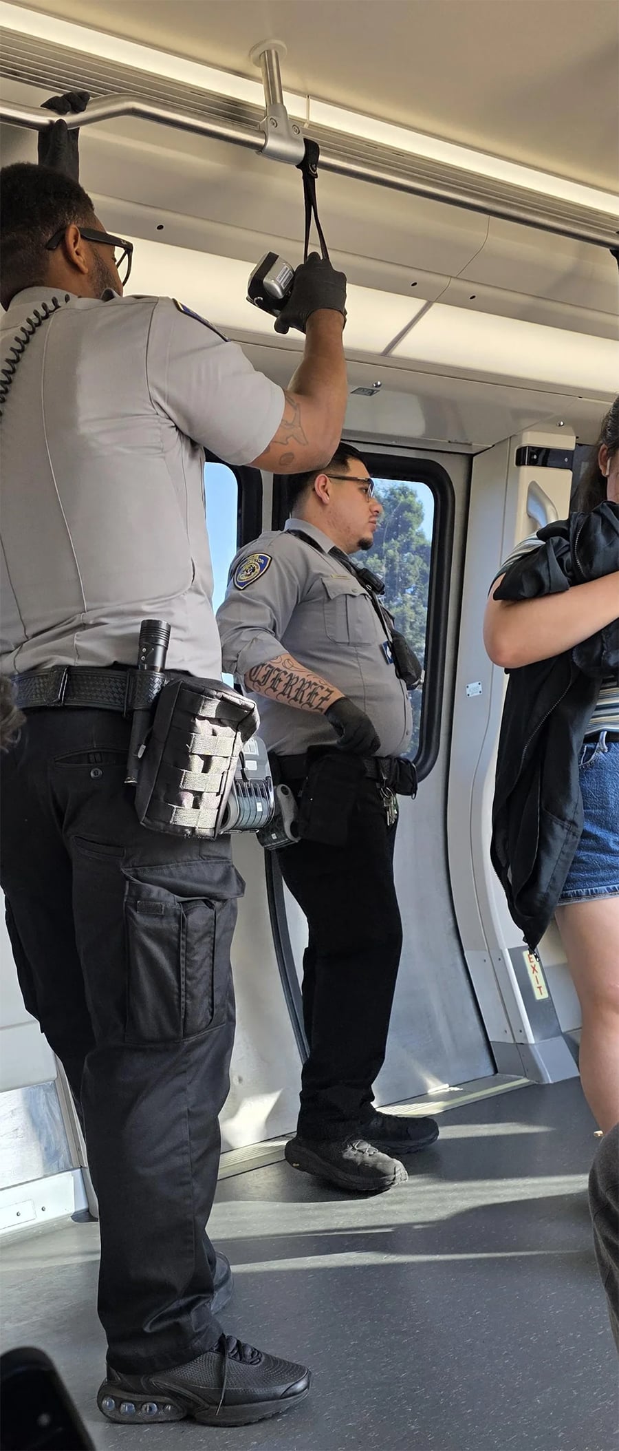 Two uniformed security officers stand inside a train. One holds an overhead rail, both wear utility belts with gear, and another person in casual clothes stands nearby. Sunlight streams through the windows.