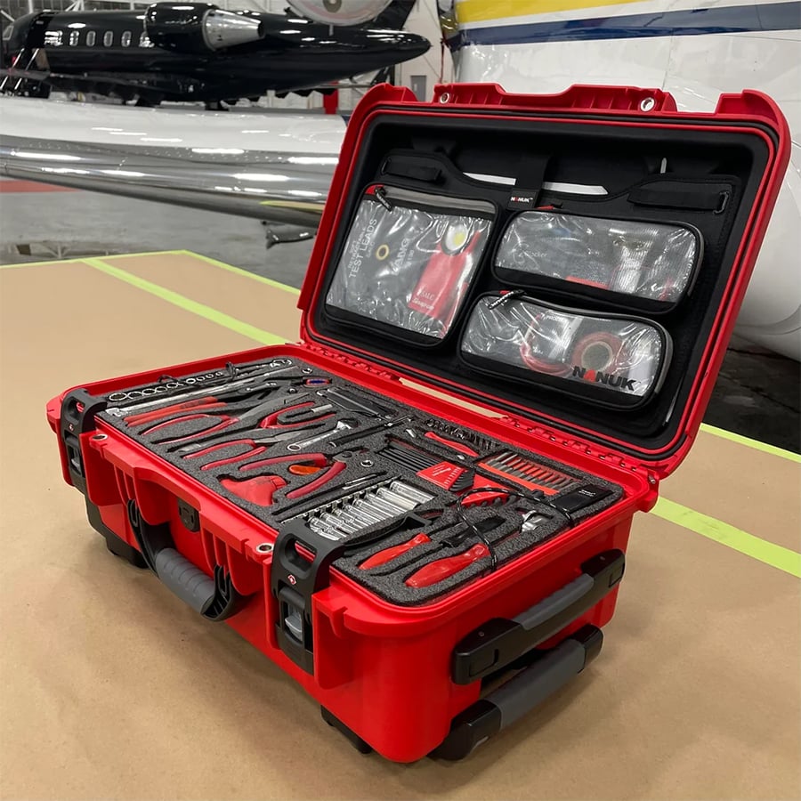 A red hard case containing various hand tools neatly organized in foam, with wrenches, pliers, and screwdrivers. Behind the case are two aircraft in a hangar.