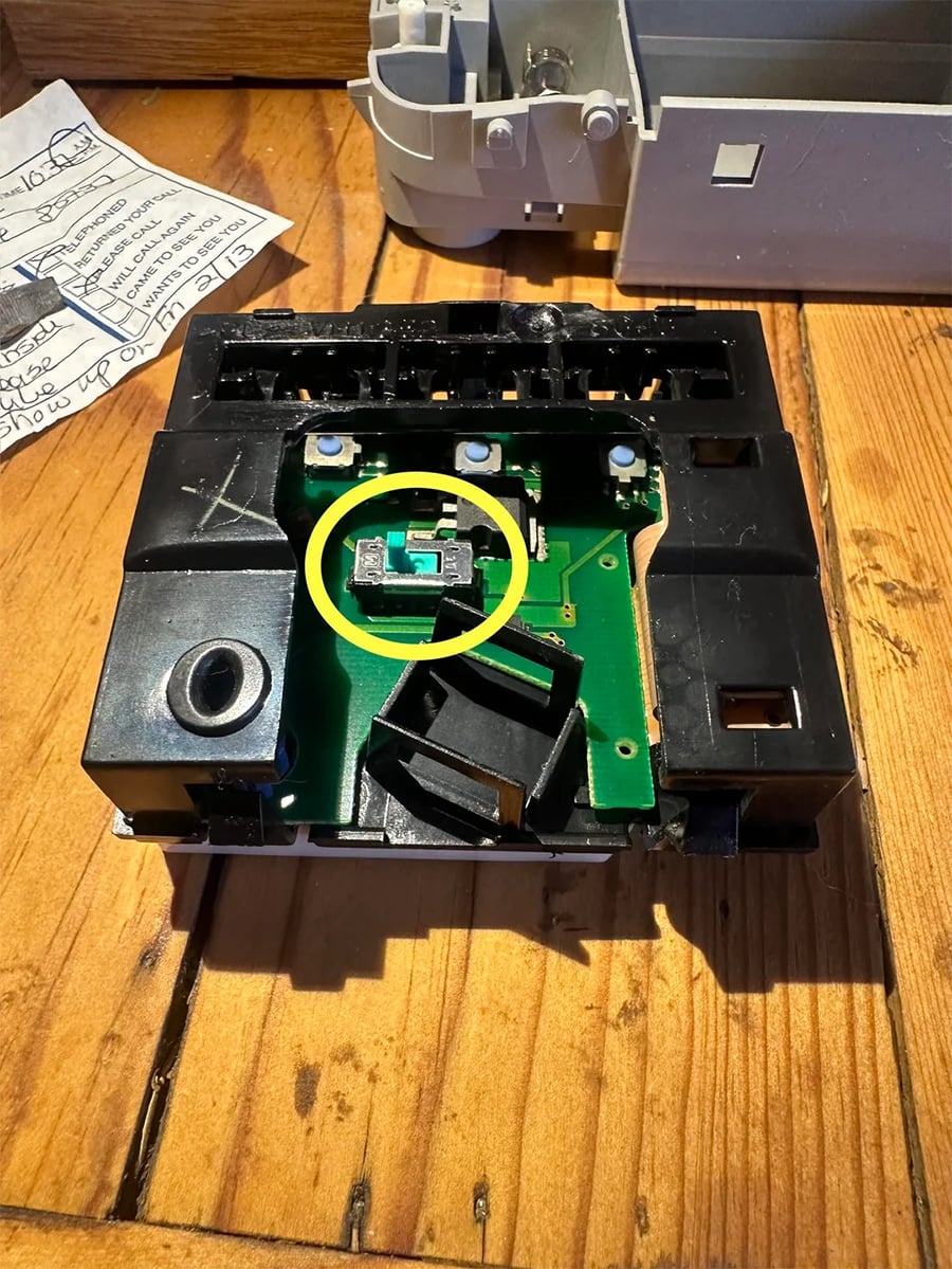 An opened electronic device with a green circuit board inside. A yellow circle highlights a small component in the center, surrounded by black plastic parts. Handwritten notes and another device are in the background.