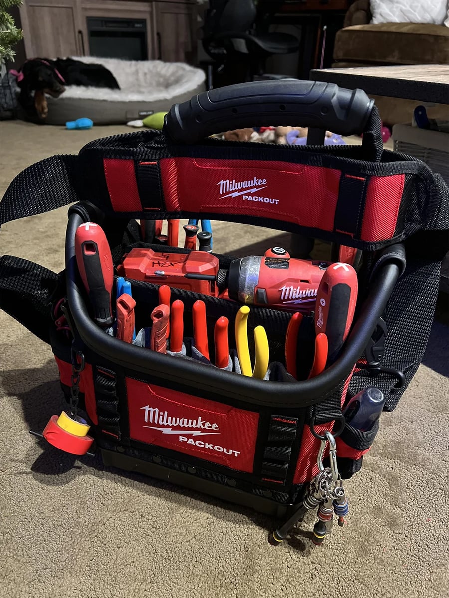 A Milwaukee Packout tool bag sits on the floor, filled with various hand tools like screwdrivers, pliers, tape measure, and a drill. Keys are clipped to the bag’s handle, and furniture is visible in the background.