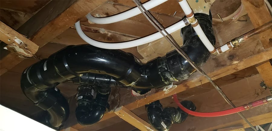 Exposed basement ceiling showing black PVC plumbing pipes and white and red PEX water lines attached to wooden joists.