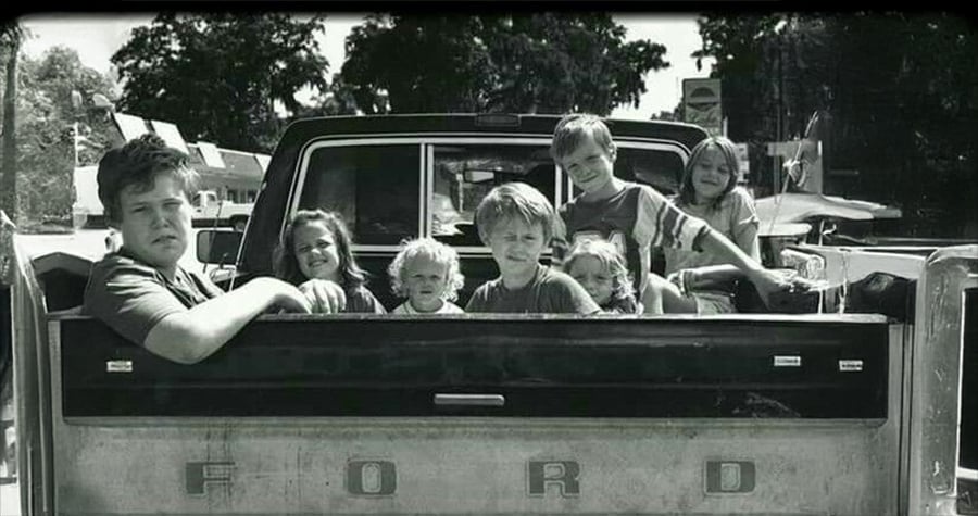 Seven children sit in the bed of an old Ford pickup truck, smiling and looking at the camera. The scene is outdoors on a sunny day with trees and buildings in the background. The photo is in black and white.