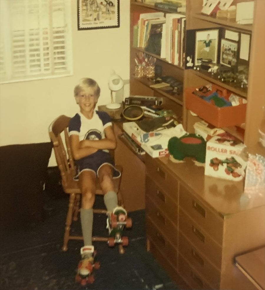 A smiling child with blonde hair sits on a chair in a bedroom, wearing roller skates and a blue outfit. The room has a desk, bookshelves with toys, and various items neatly arranged.
