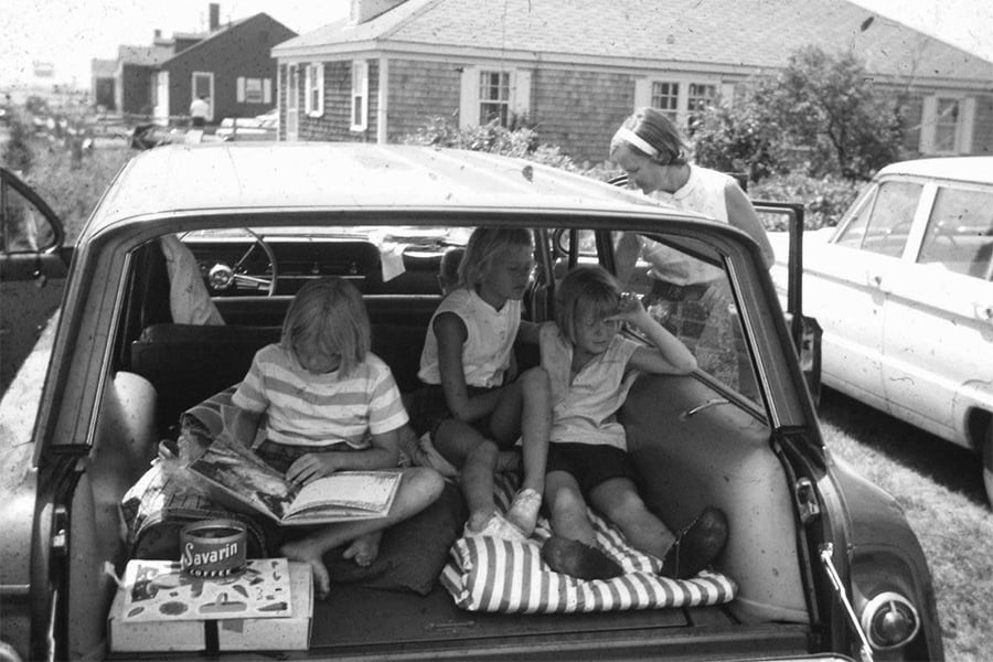 Three children sit in the back of an open station wagon, reading and relaxing. A woman stands nearby, looking inside. Houses and a parked car are visible in the background on a sunny day.