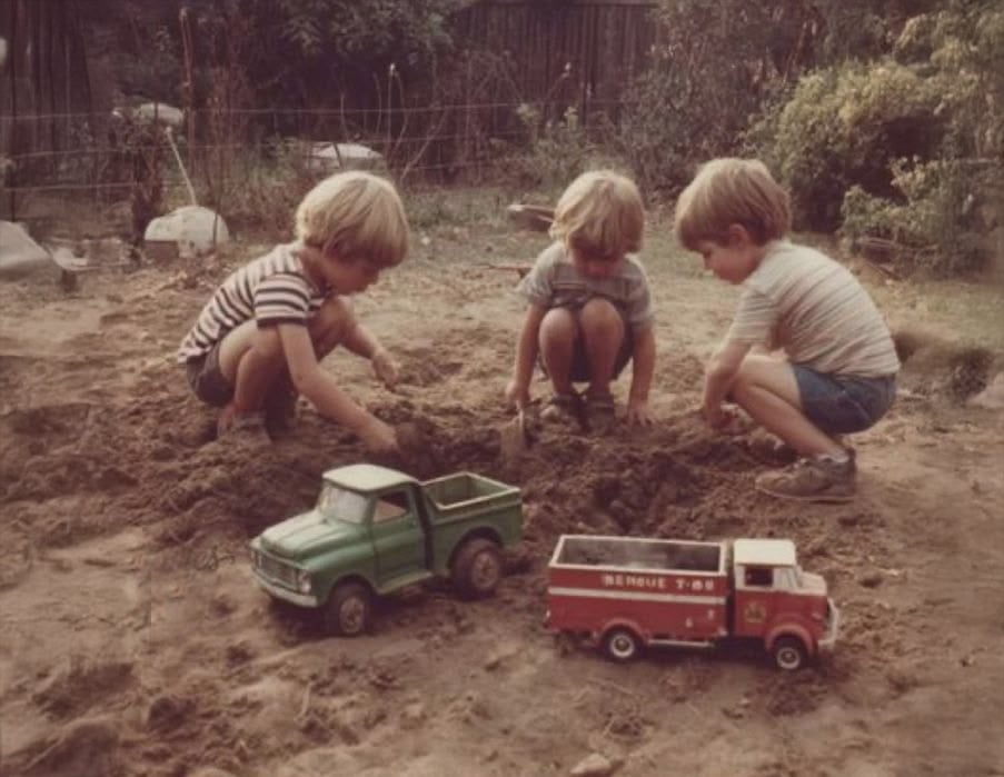 Three young children play in the dirt with toy vehicles, including a green truck and a red fire engine, in a sunny outdoor yard surrounded by greenery and a wire fence.