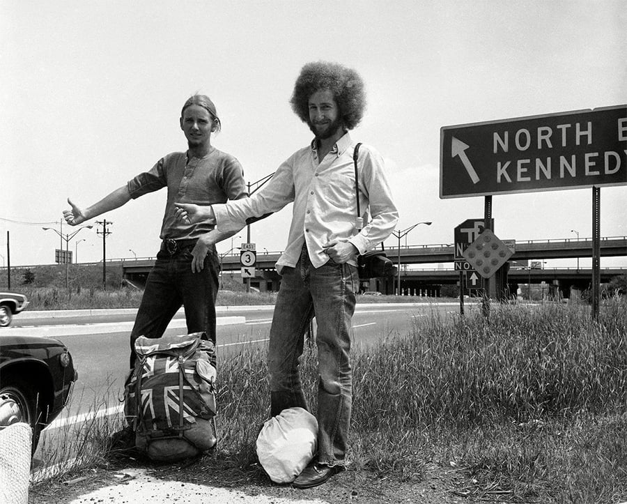 Two young men hitchhike by a roadside, holding out their thumbs. They stand beside backpacks, one with a British flag. A highway sign points toward “North East Kennedy.” An overpass and a car are visible in the background.