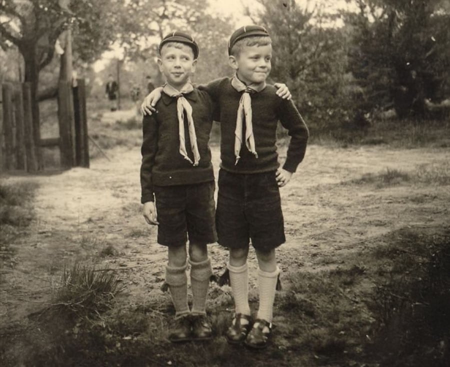 Two young boys in matching uniforms and hats stand outdoors, arms around each other, smiling. They wear shorts, sweaters, neckerchiefs, long socks, and shoes. Trees and a fence are visible in the background.