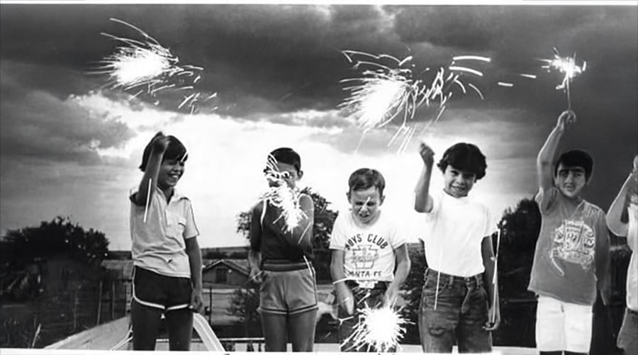 Five children stand outside at dusk, smiling and holding sparklers in the air, with trees and houses in the background under a cloudy sky.