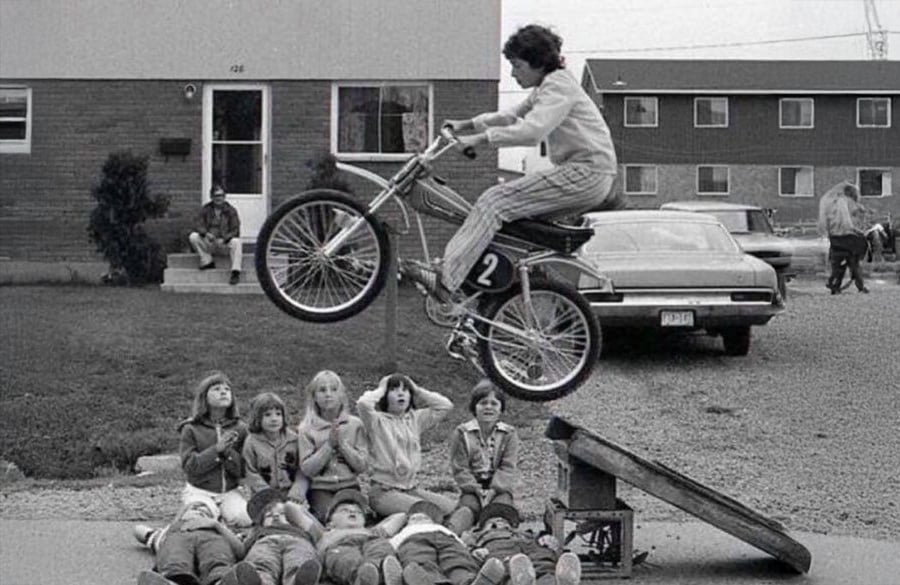 A boy on a bike jumps over a ramp above several children lying side-by-side on the ground, with houses and cars in the background.