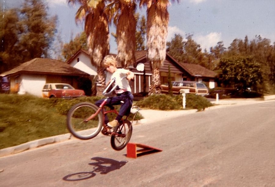 A person on a BMX bike performs a jump off a small ramp on a suburban street with palm trees and houses in the background.