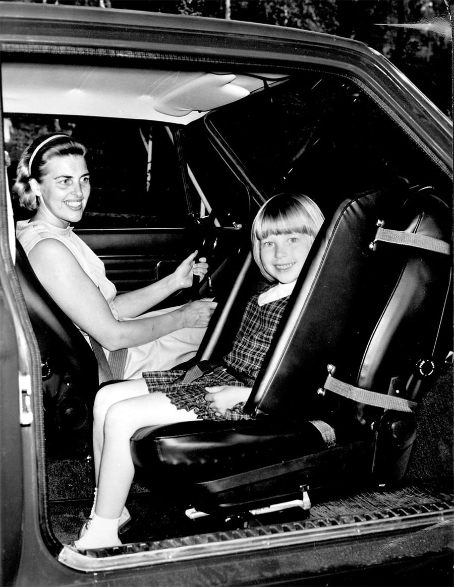 A woman smiles while sitting in the driver’s seat of a car, and a young girl with short blonde hair smiles in the back seat, both looking at the camera.