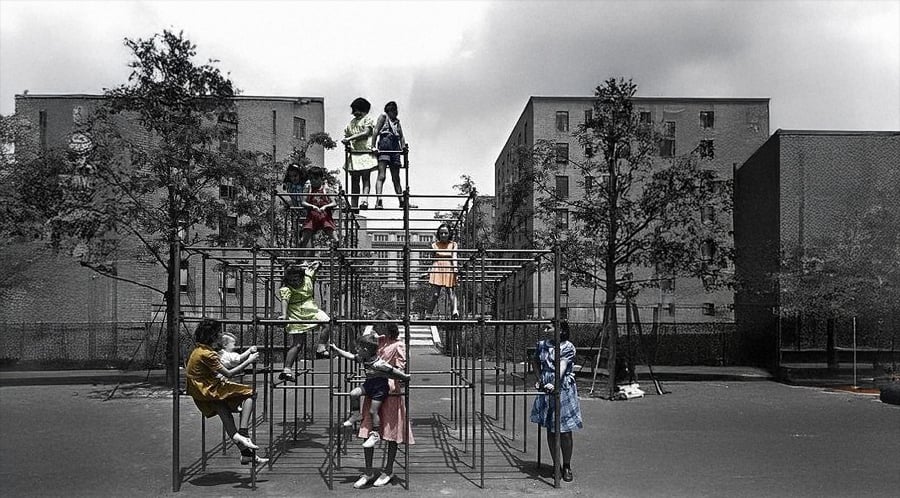 Children in colorful dresses play on a large metal jungle gym in an urban playground, surrounded by brick buildings and sparse trees under a cloudy sky.