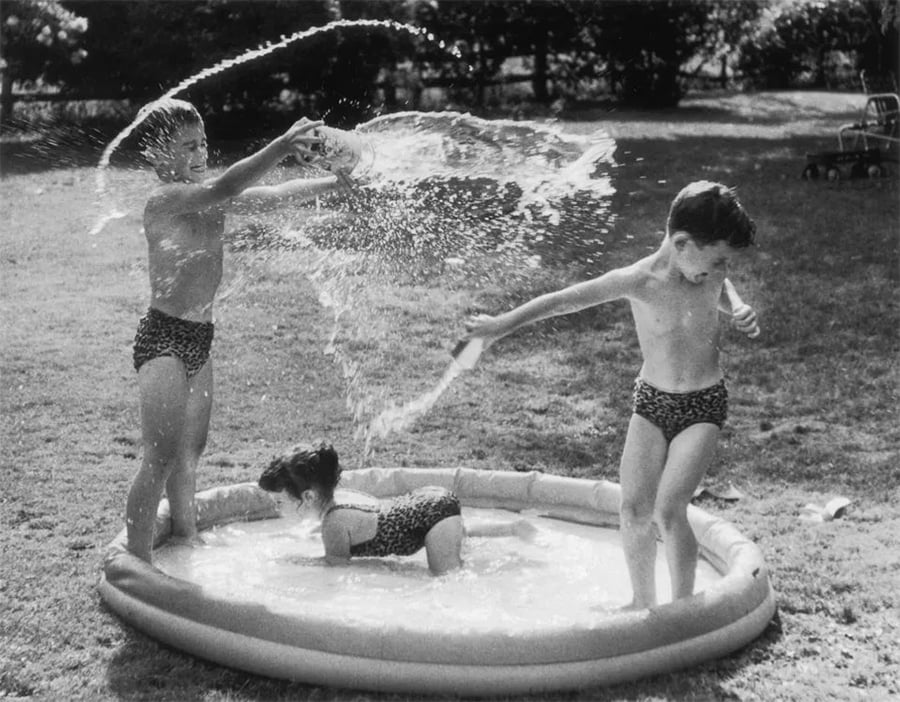 Three children in swimsuits play in a small plastic pool. One splashes water with a container, another bends over in the pool, and the third stands nearby, avoiding the splash. Grass and trees are in the background.