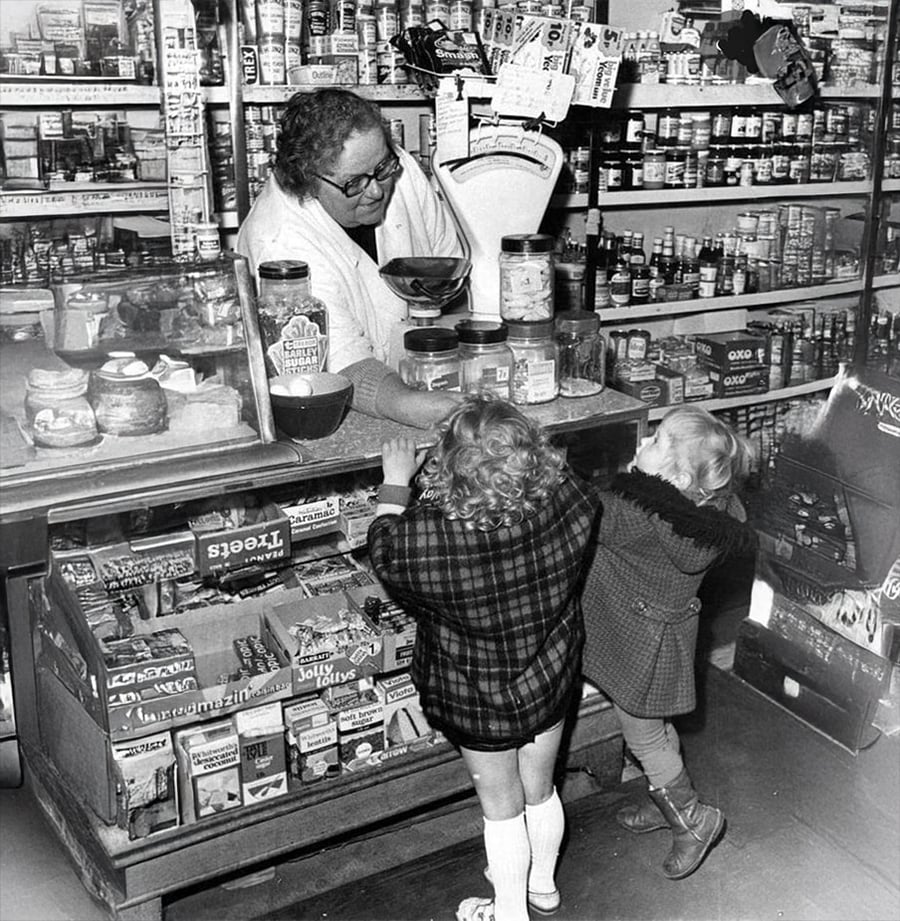 A shopkeeper behind a counter weighs items on a scale while two young children in coats stand in front, looking up. The counter displays jars, snacks, and various goods, with shelves of canned food in the background.