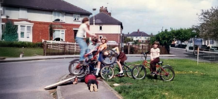 Children play on bikes near a small ramp on a suburban street. One child is mid-jump on a bike, others watch, and one child lies face-down on the sidewalk. Houses and trees are in the background.