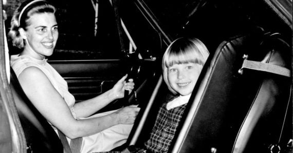 A woman sits in the driver’s seat of a car, smiling, while a young girl with blond hair and bangs sits in the front passenger seat, also smiling.