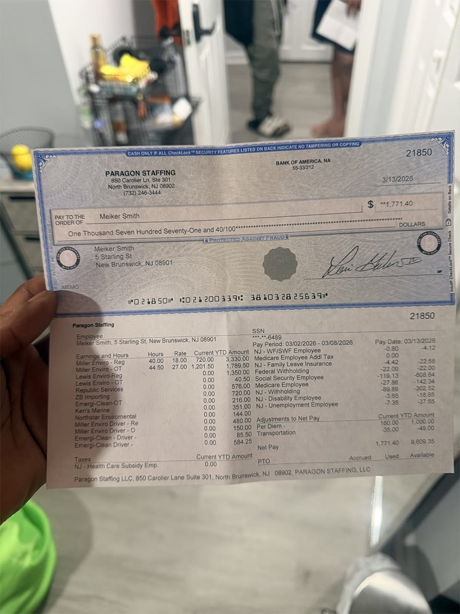 A hand holds a paycheck and pay stub from Paragon Staffing, showing payment details, deductions, and a net pay of $1,771.40. Background includes a kitchen counter with fruit, and a person’s reflection in a mirror.