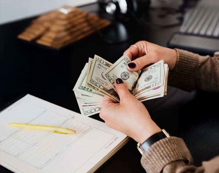 A person with dark nail polish counts U.S. dollar bills at a desk with a yellow pen, a notebook, and a computer keyboard in the background.