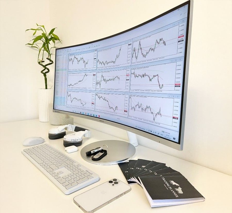 A modern desk with a curved monitor displaying multiple financial charts, a keyboard, mouse, notepads, a smartphone, and a small potted plant.