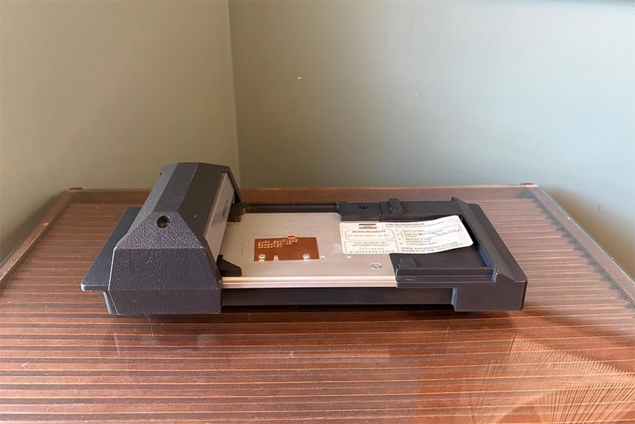 A vintage computer peripheral with a black plastic casing and metal base sits on a wood surface against a light green wall. Labels and a circuit are visible on the device.