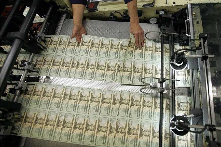 A person handles sheets of newly printed U.S. hundred dollar bills coming out of a large printing machine in a currency production facility.