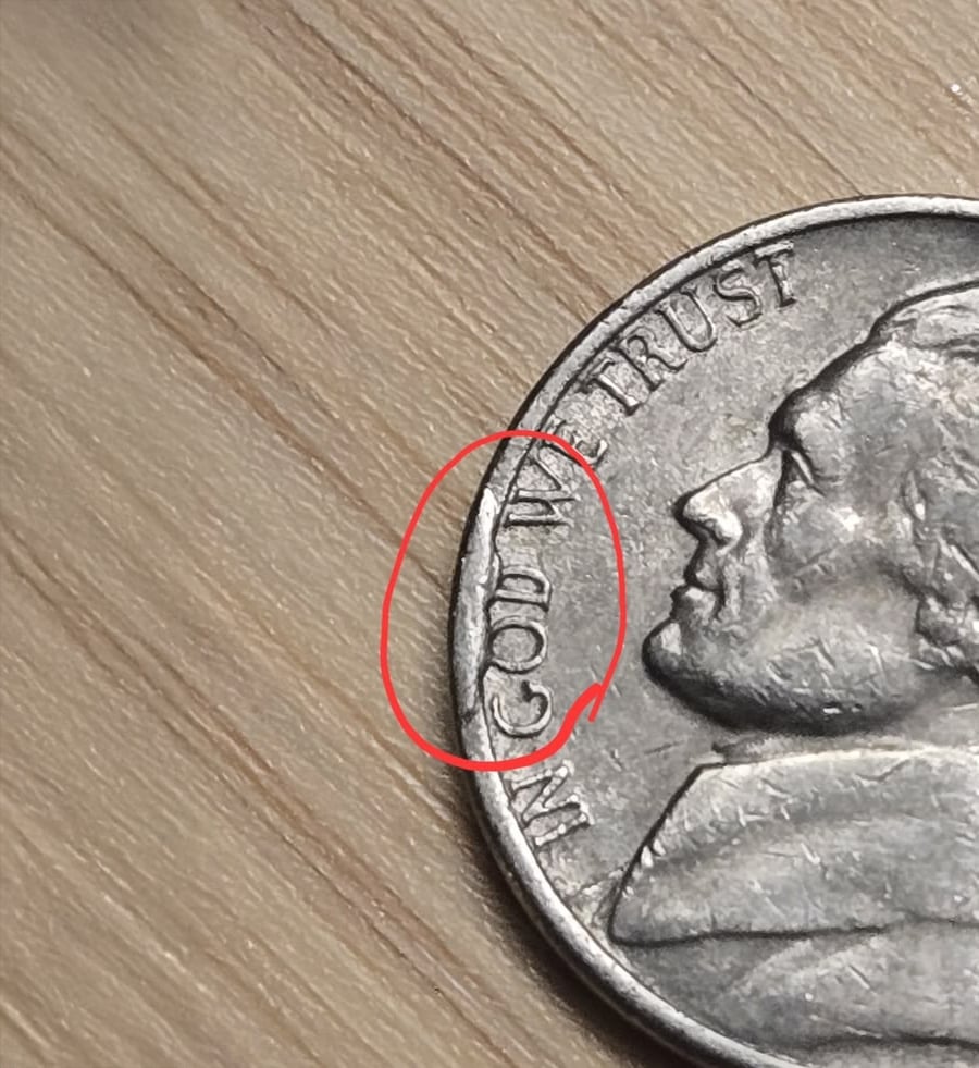 A close-up of a U.S. nickel with part of the phrase “IN GOD WE TRUST” circled in red on the coin’s left edge, lying on a wood surface.