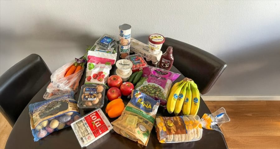 A table with groceries including bananas, apples, carrots, tofu, mushrooms, bread, salad greens, pasta, bell pepper, berries, yogurt, chocolate syrup, canned beans, and grape tomatoes.