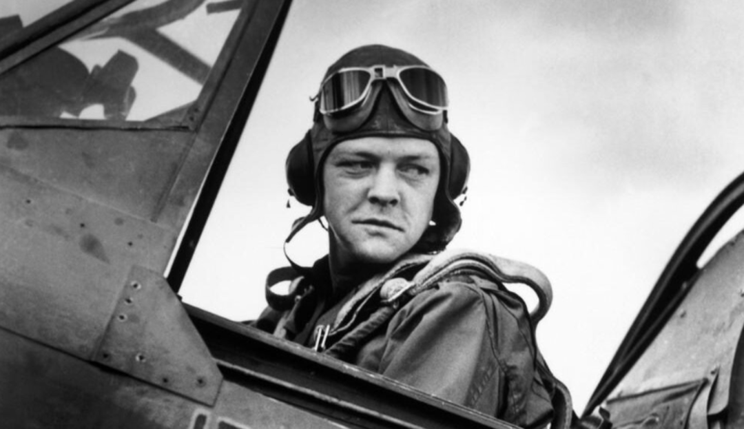 A World War II pilot wearing a flight helmet and goggles sits in the cockpit of a military aircraft, looking off to the side.