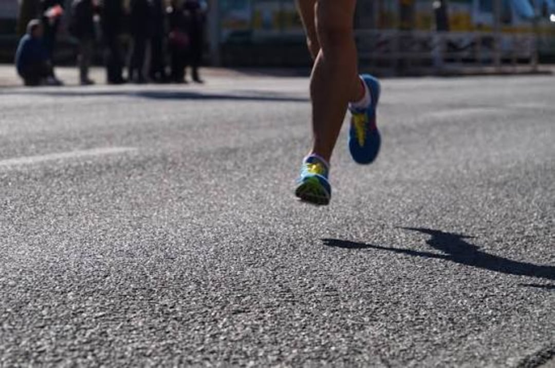 Runner’s legs and colorful shoes in motion on a sunlit asphalt road, with a shadow cast on the ground and blurred figures gathered in the background.