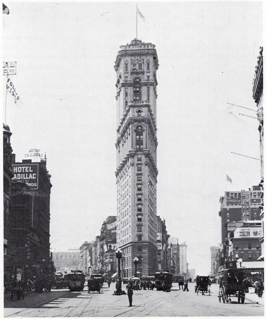 A historic black-and-white photo of a tall, narrow flatiron-style building on a busy city street with horse-drawn carriages, streetcars, and pedestrians. Signs for hotels and businesses are visible.