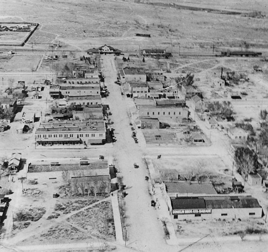 Aerial view of a small, sparsely populated town with dirt roads, scattered buildings, cars parked on streets, and open land in the background.