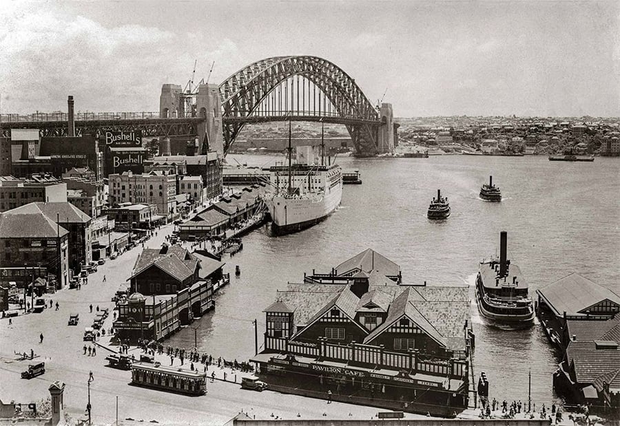 Historic photo of Sydney Harbour with ferries, a large ship docked, buildings, and the Sydney Harbour Bridge under construction in the background. Crowds of people and trams are visible along the waterfront.