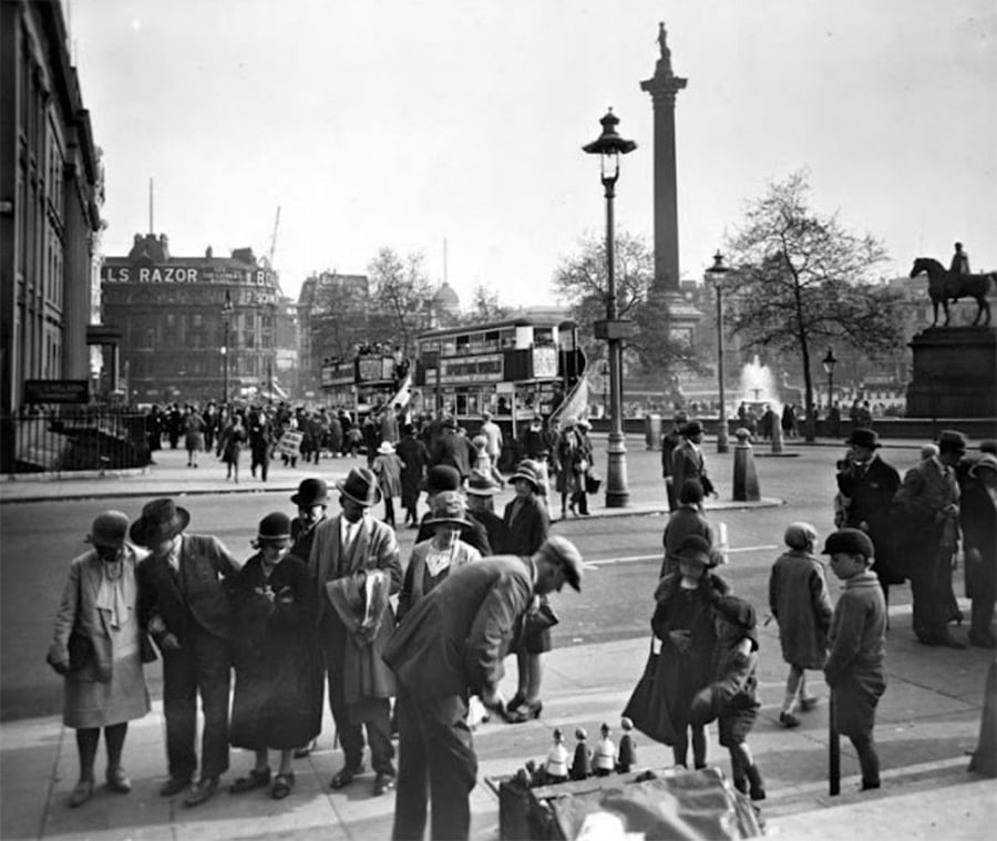 A busy street scene in London, with people gathered around a street vendor. Double-decker buses and statues are visible, including Nelson’s Column in the background. Trees and lampposts line the area under a cloudy sky.